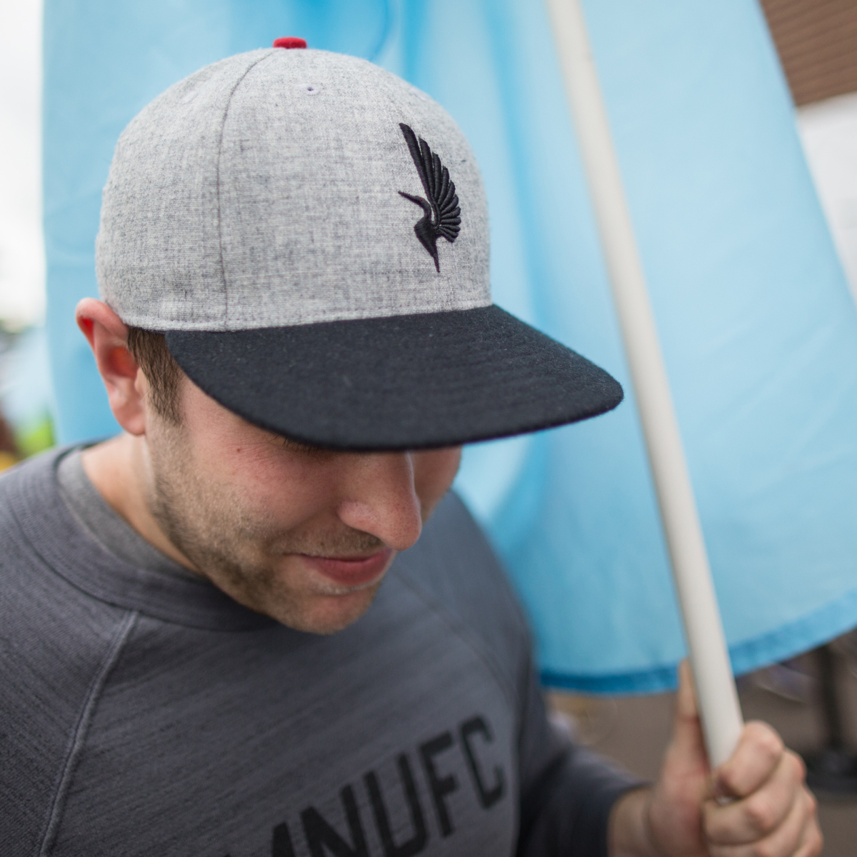 A man wearing MNUFC merch on gameday. A grey hat with the black loon from the logo. The top button on the hat is red to match the loon's eye.
