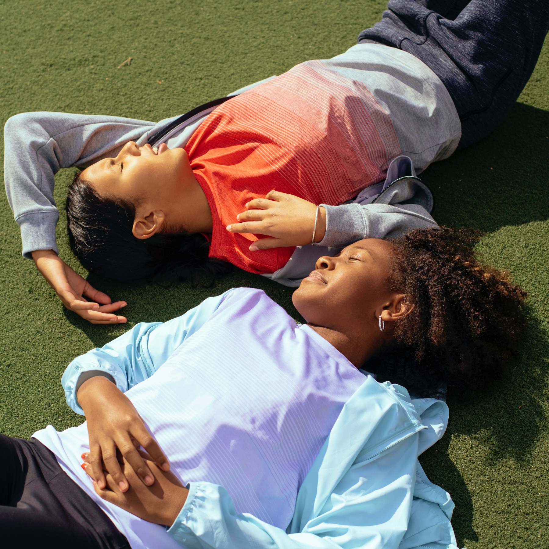 Two adolescent-age girls relaxing in the sun on a turf-like surface, with their eyes closed.