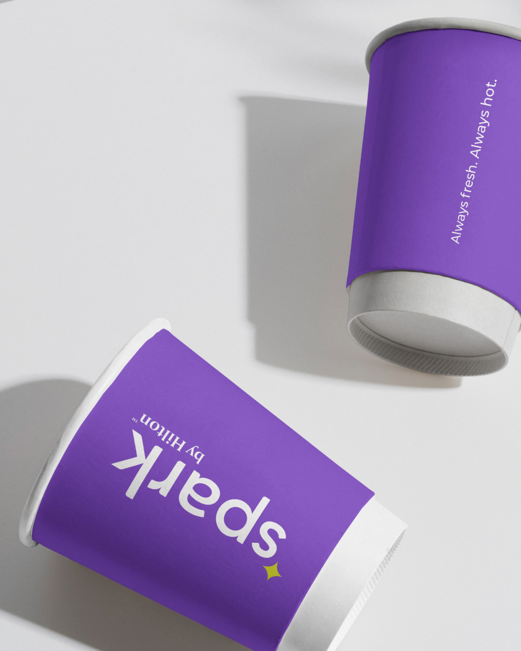 Two purple disposable coffee cups laid down on a white surface, with logo and white text that reads, “Always fresh. Always hot.”