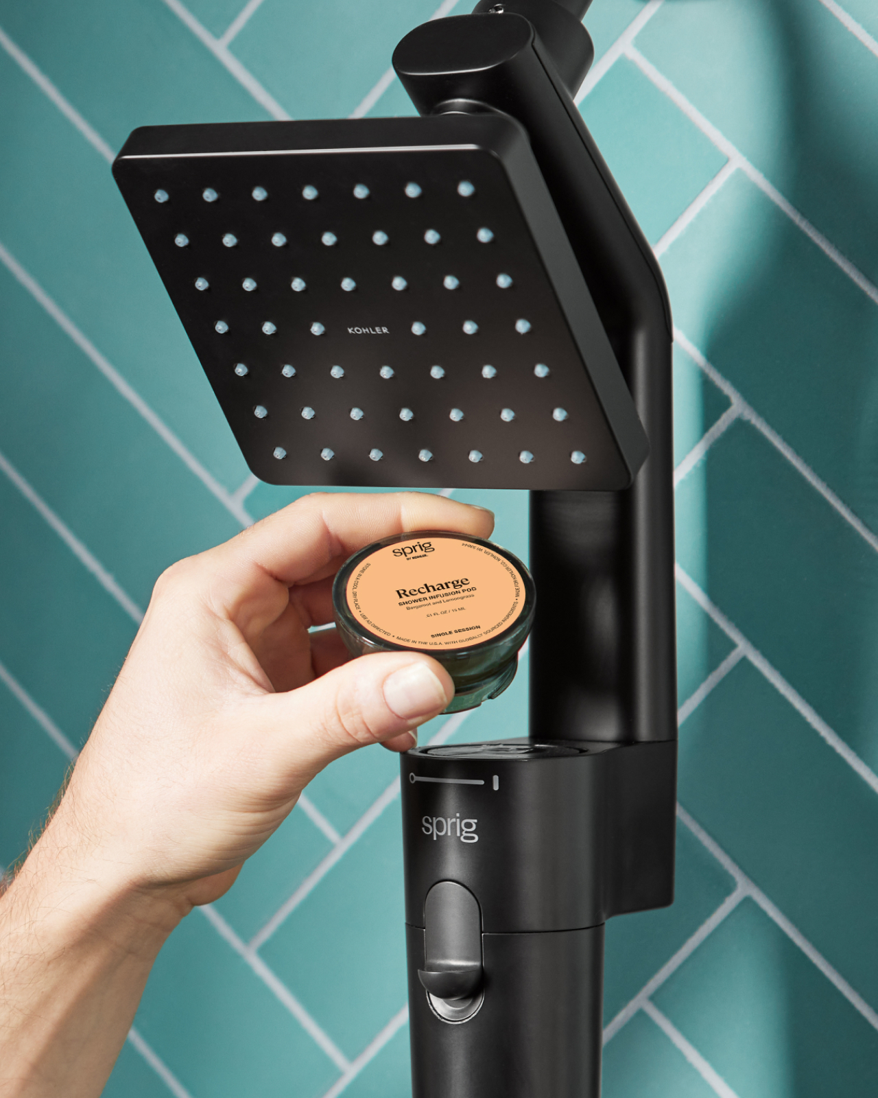 Close-up of a man’s hand inserting a “Recharge”-scented Shower Infusion Pod into a Sprig Shower Infusion showerhead, with blue tile behind.