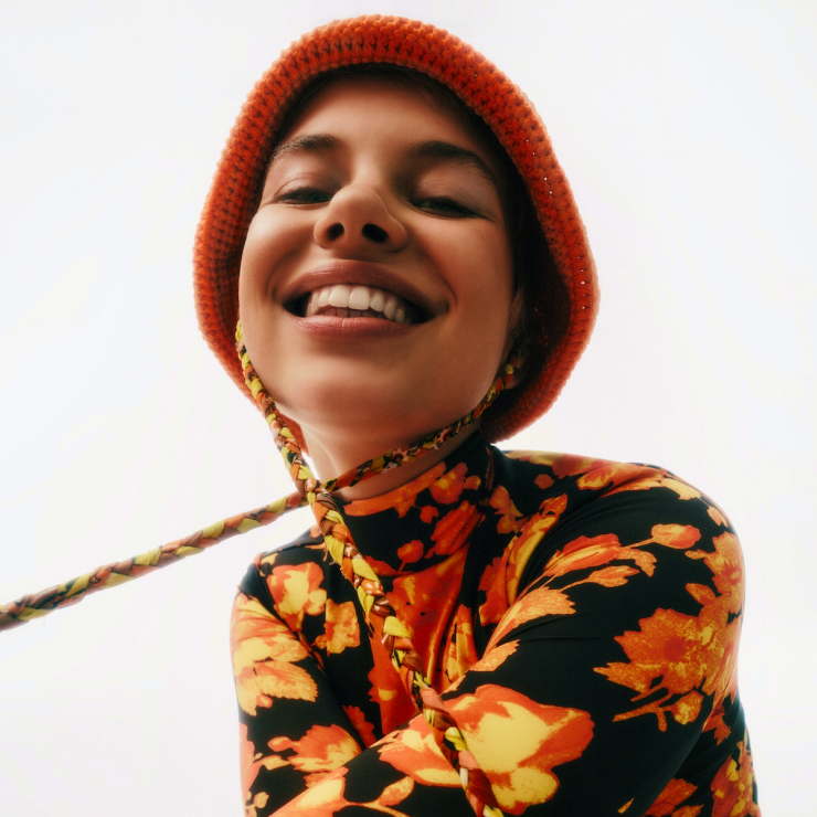 Headshot of a young woman wearing makeup, dressed in a floral shirt and orange hat, and smiling.