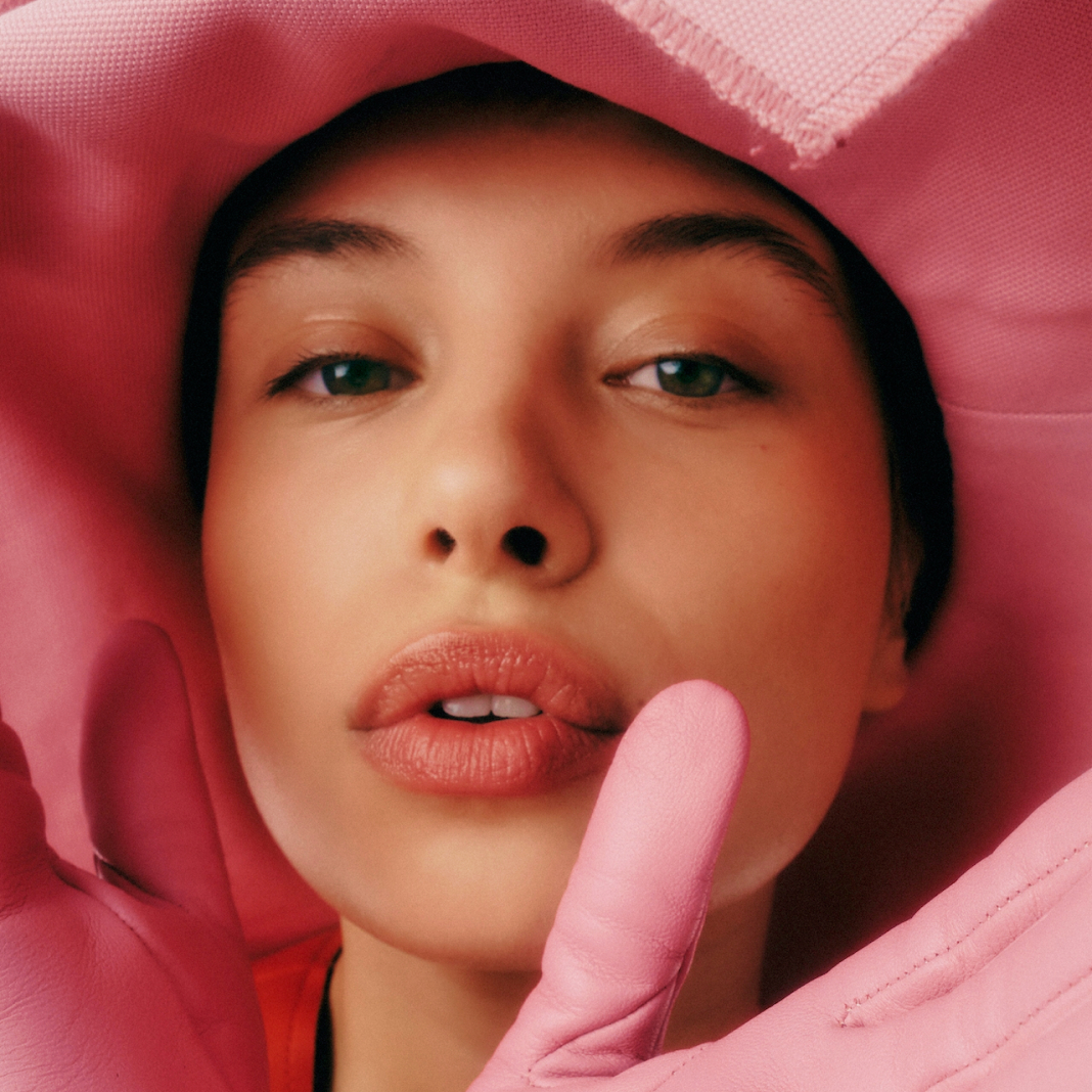 Closeup on the face of a young woman wearing a pink hat and gloves, showing her smooth skin and expertly applied makeup.